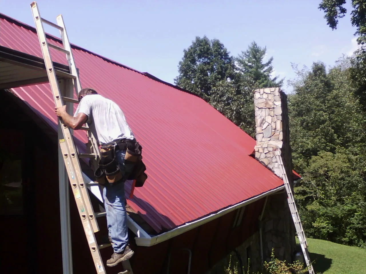 Roofer on a ladder beside a red metal roof and stone chimney — Soffit & Fascia Repair by Roofing Brothers in Beaver